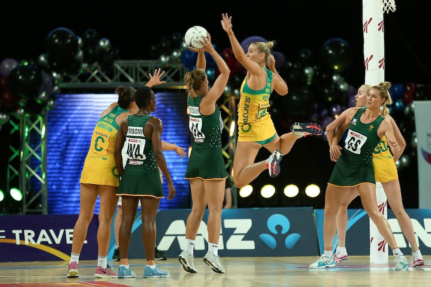 Courtney Bruce leaps with her left hand over the ball and both feet bent back in the air. She is wearing a green and gold dress.