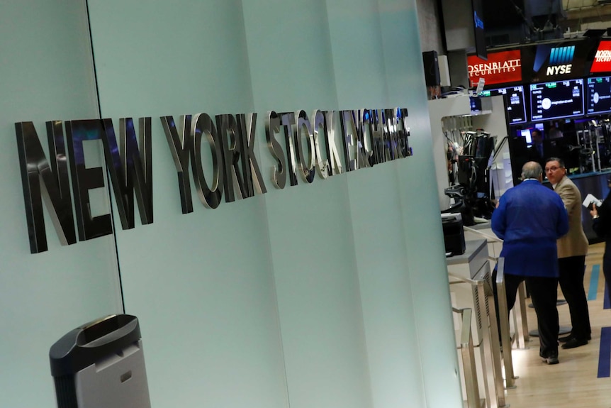 A shiny silver sign along the wall of a modern building, with two men talking on trading room floor.