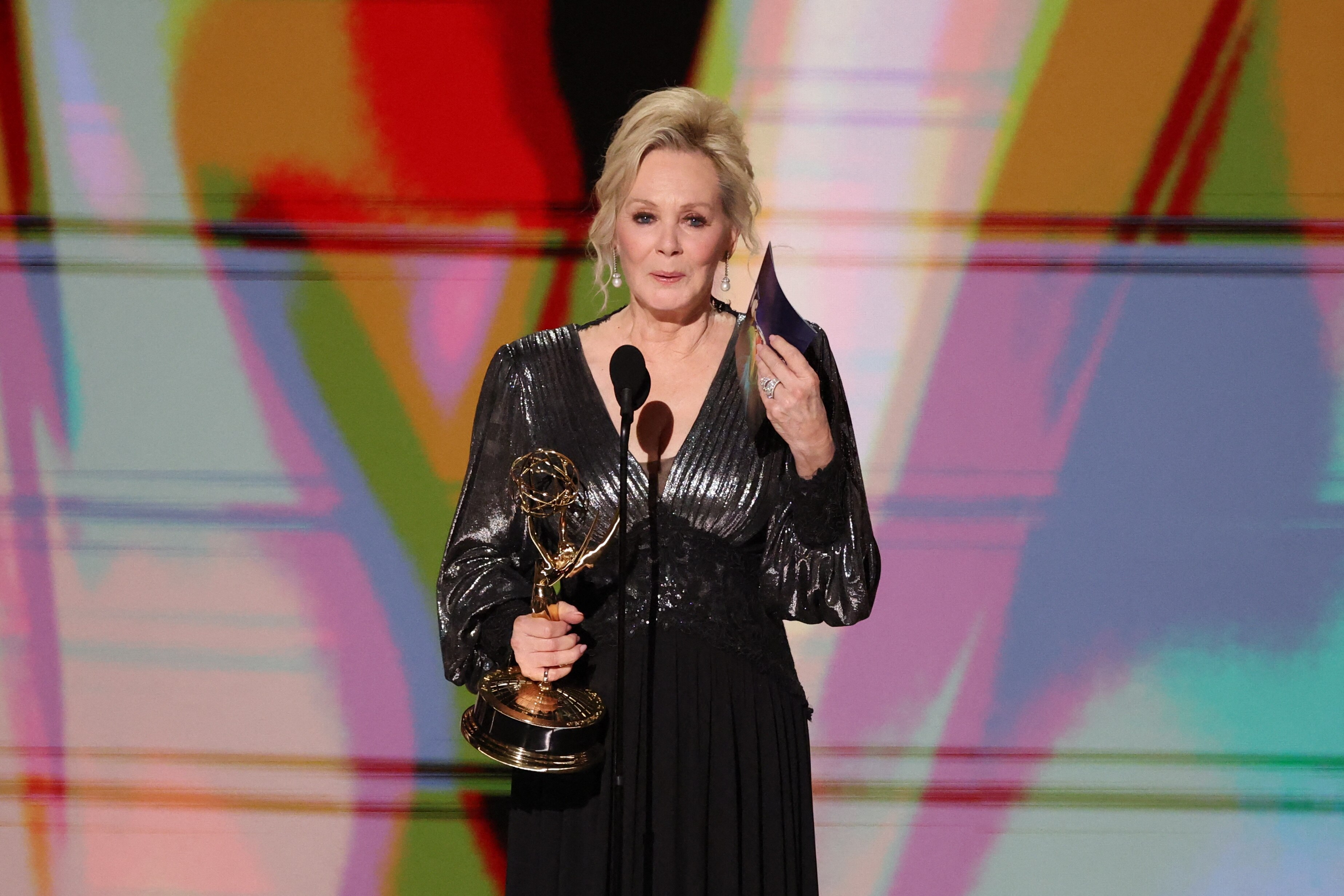 Jean Smart in black dress holding Emmy waves back tears on stage.