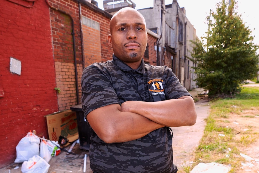 A man stands on a Baltimore sidewalk with his arms folded over his chest