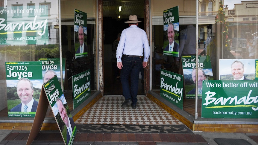 Barnaby Joyce has his back to the camera as he walks into his office with campaign posters stuck to the windows.
