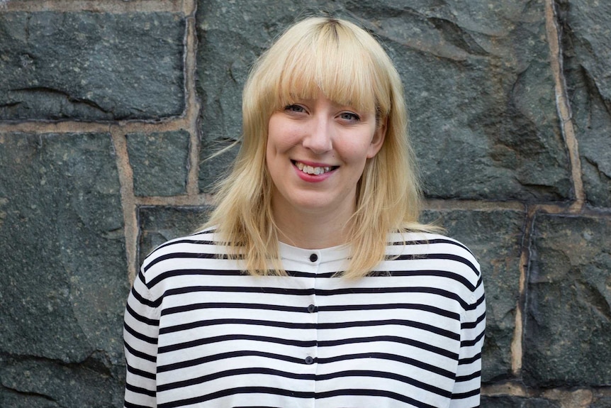 Young blonde woman in striped top sitting in front of a stone wall.