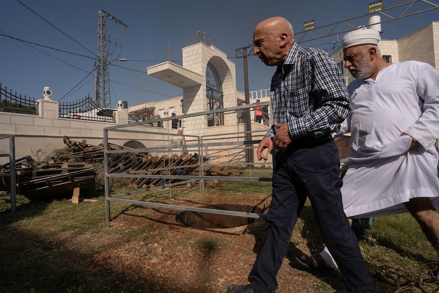 Two men walk around an empty pit with stacks of firewood in the background