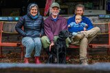 An older couple with their adult son and baby girl sitting on a bench sea with their black labrador dog