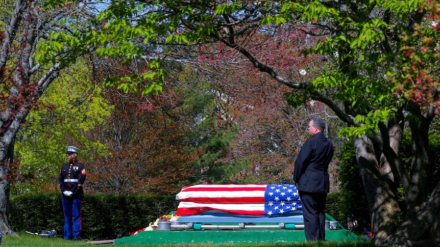 A coffin draped in a US flag sits in a clearing, with a man in a suit and a US marine in full dress uniform nearby