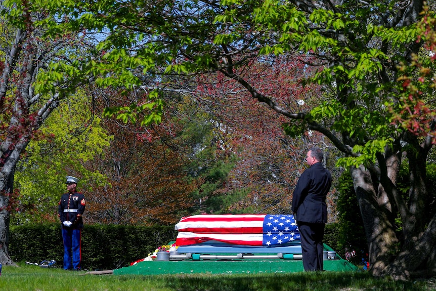 A coffin draped in a US flag sits in a clearing, with a man in a suit and a US marine in full dress uniform nearby