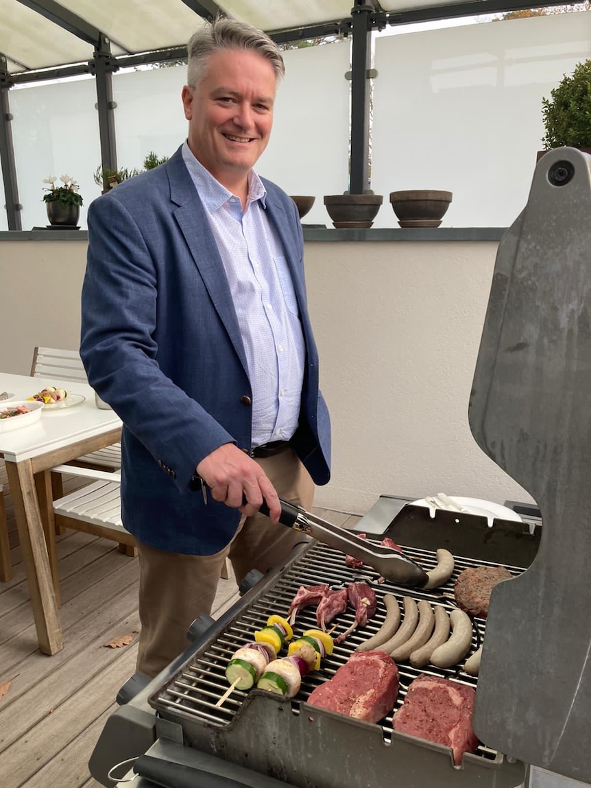Mathias Cormann holds tongs while standing at a barbecue with sausages and steaks