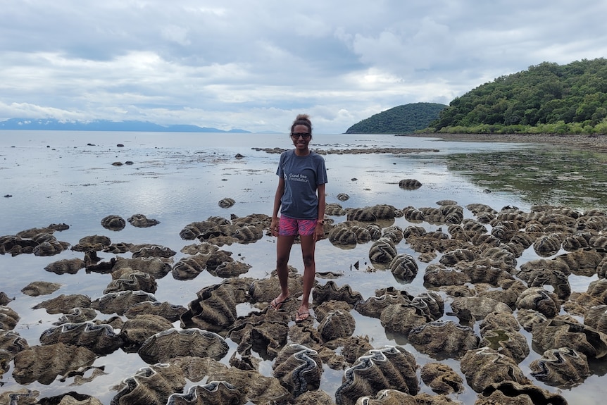 A woman stands near some giant clams with the ocean and a hill in the background.