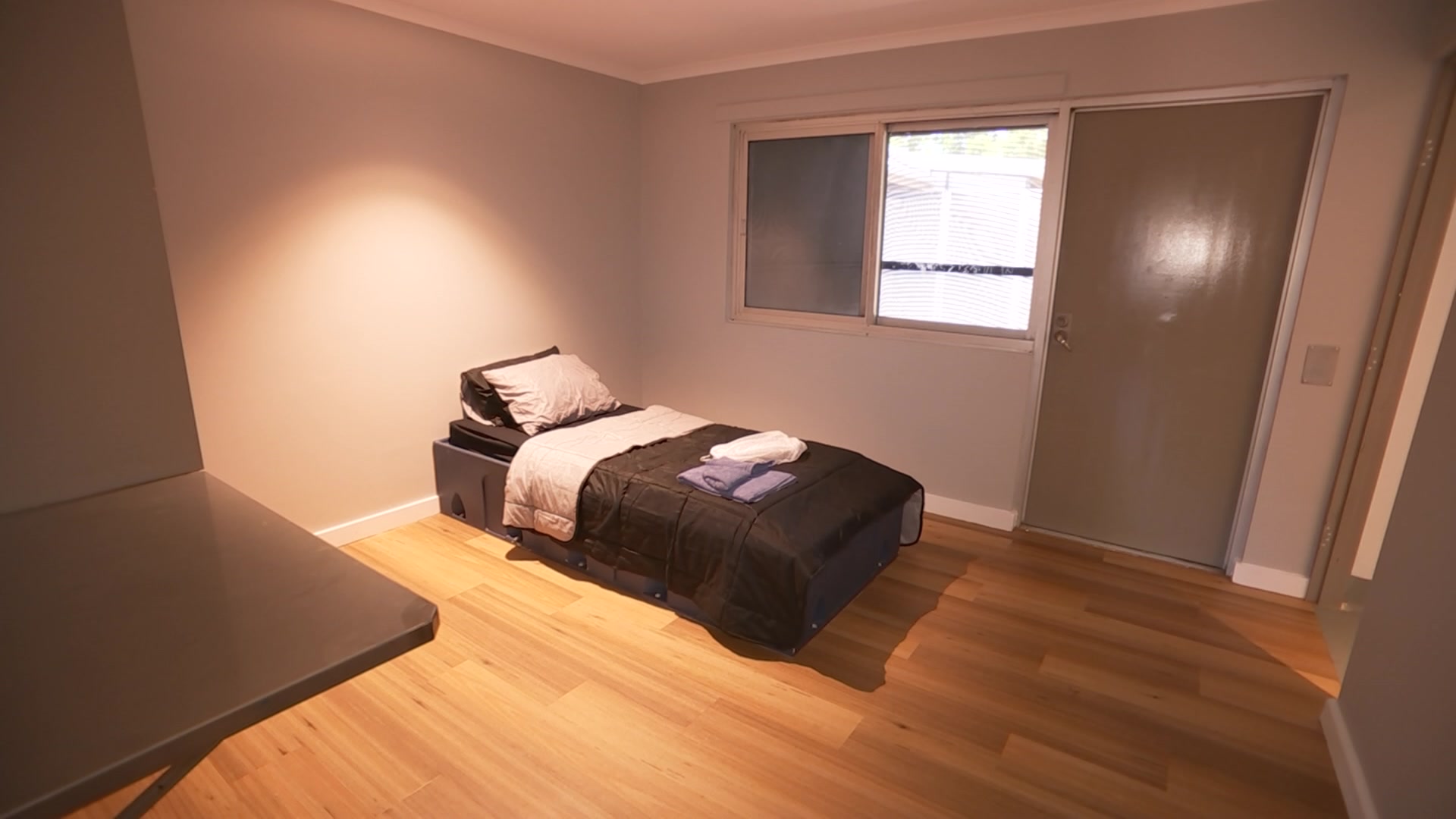 Um quarto vazio, piso laminado de madeira, cama de solteiro com toalhas e roupas na cama, e uma pequena janela e porta.