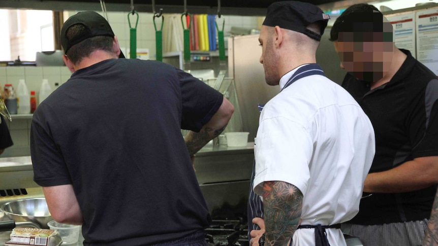 A man in a chef's uniform supervising two other men in a commercial kitchen.