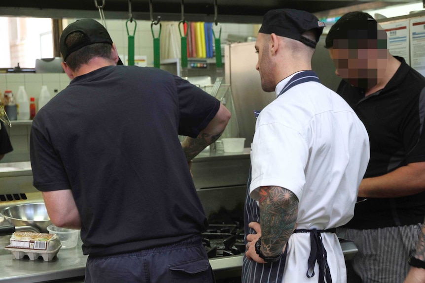 A man in a chef's uniform supervising two other men in a commercial kitchen.