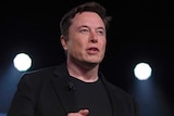 Elon Musk wears a black t-shirt and jacket and lifts his hands.