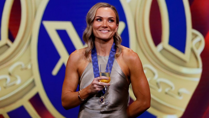 Emily Bates smiles in front of the AFLW Best and Fairest signage with a glass of champagne and her medal