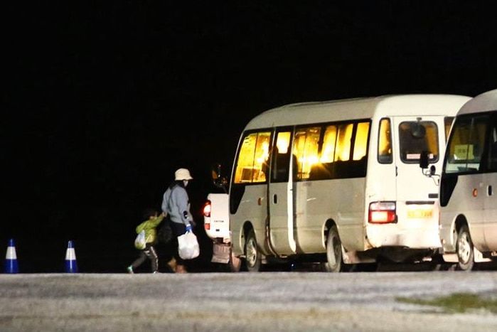 A long shot of a woman and a young boy walking towards a white bus at night with another bus behind it.