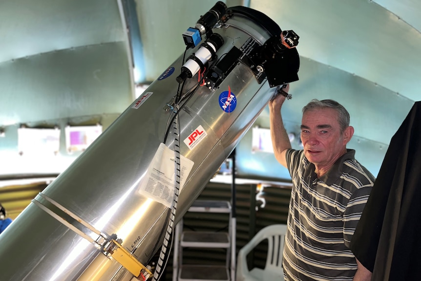 A man wearing a polo shirt standing in a dome shaped room with a hand on a large metal telescope