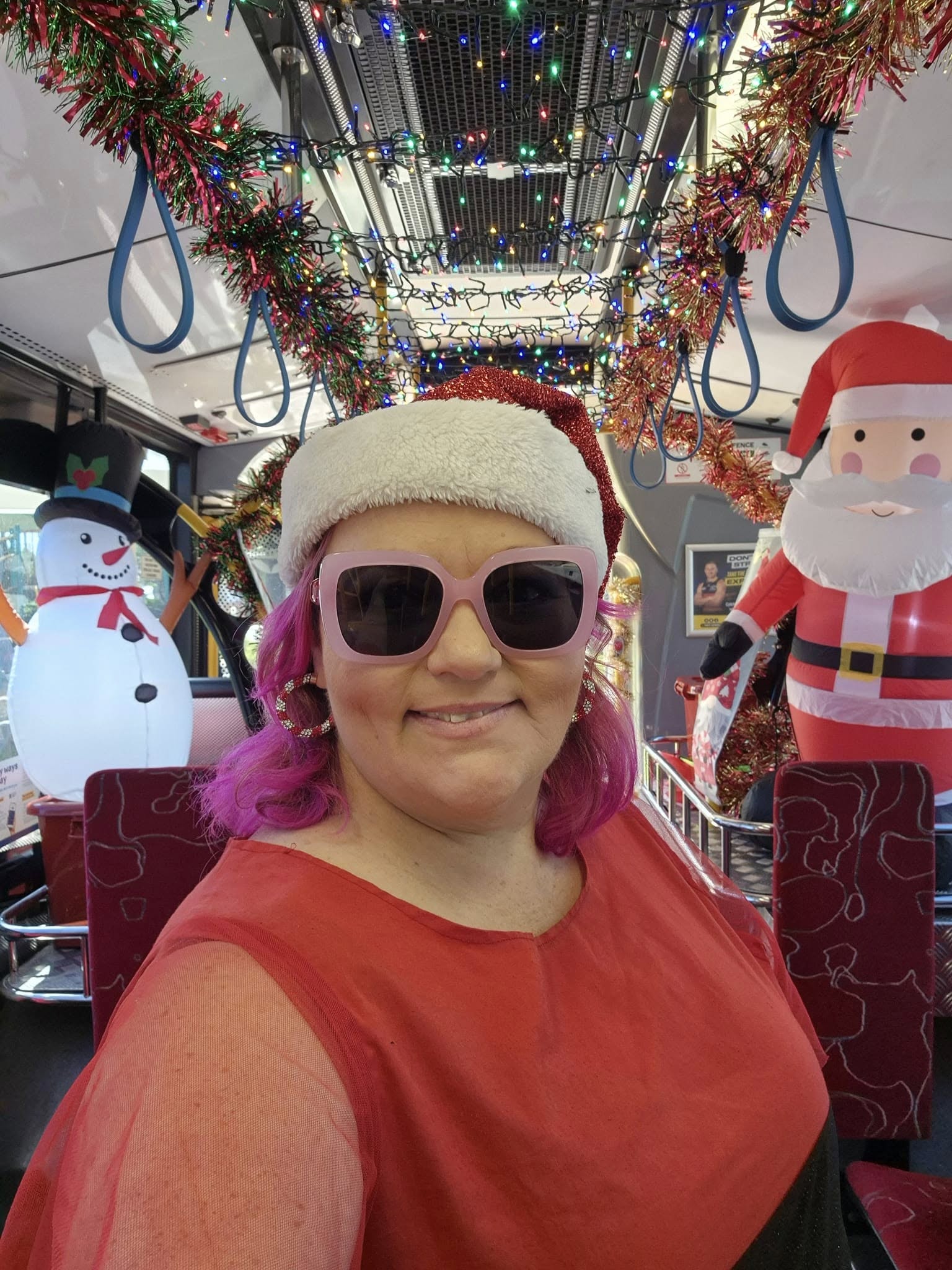 selfie of a woman in sunglasses wearing a santa hat with inflatable snowman and santa in the background on a bus