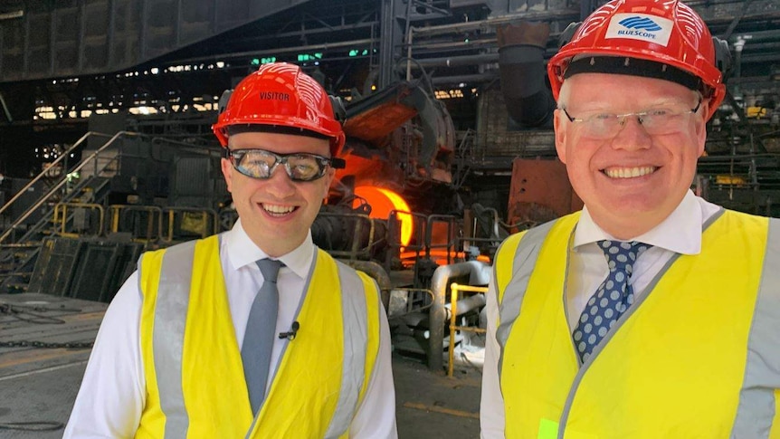 Two men stand in hard hats in a steelworks.