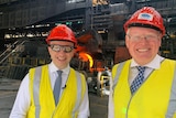 Two men stand in hard hats in a steelworks.