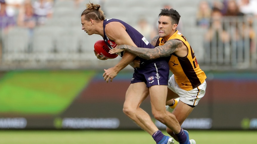 A Fremantle Dockers AFL player holds the ball as he is tackled by a Hawthorn opponent.