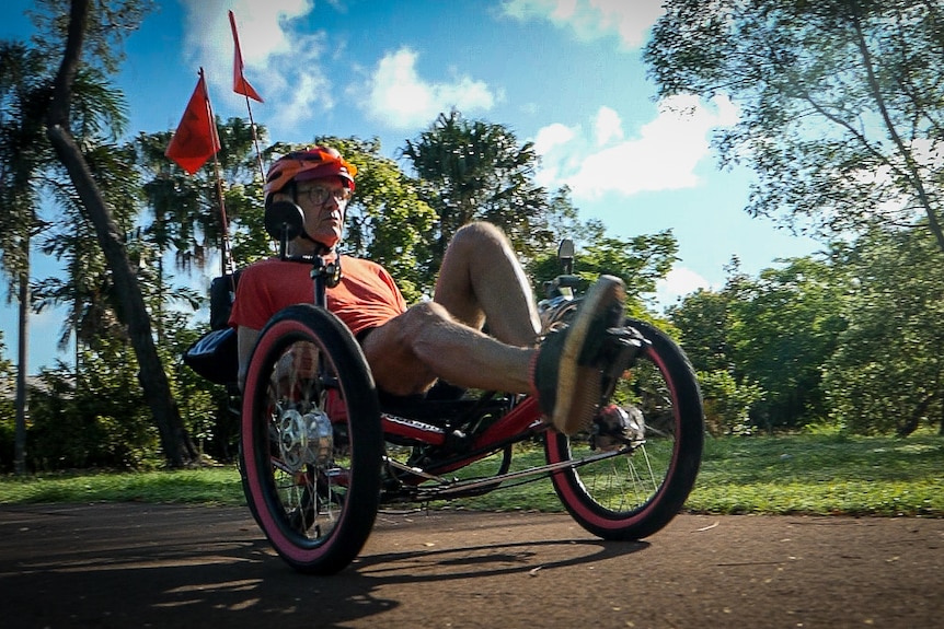 Man in bright shirt rides recumbent tricycle through a park in the early morning.
