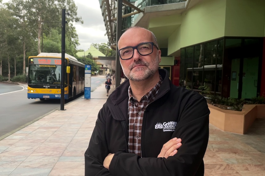 A man with glasses stands on the street with a bus in the background.