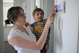 A woman is wearing glasses and looks at her air-conditioning controller on the wall with her young son on her hip.