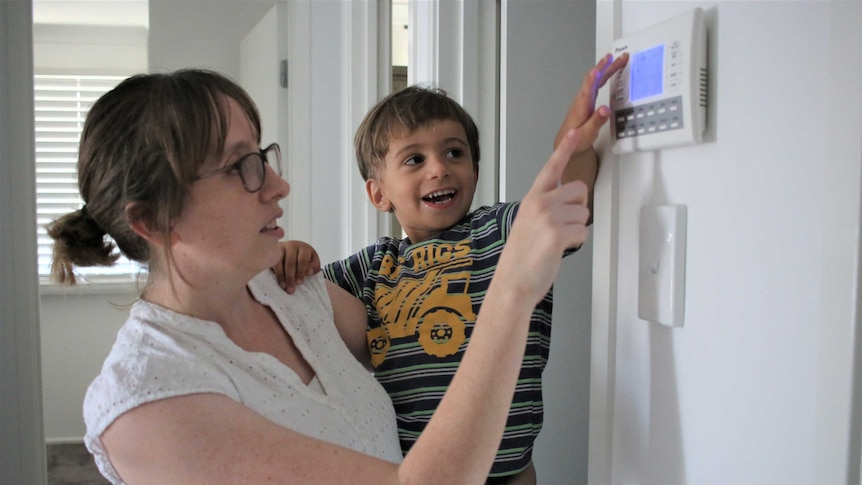 A woman is wearing glasses and looks at her air-conditioning controller on the wall with her young son on her hip.
