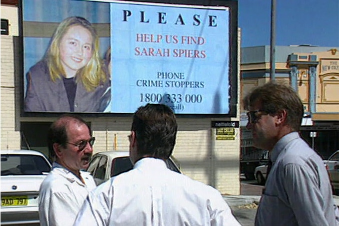 Three men stand talking in front of a billboard appealing for information about missing teenager Sarah Spiers.