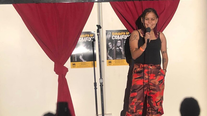 A young indigenous woman stands up on stage with a microphone with a big smile on her face.