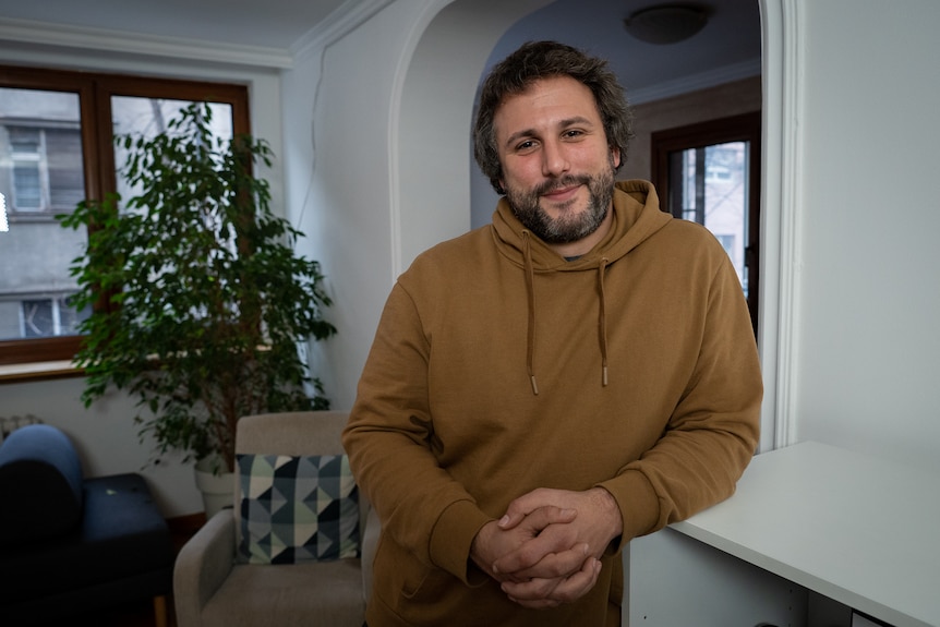 A man in a brown hoodie leans his arm on a shelving unit while posing for a photo.