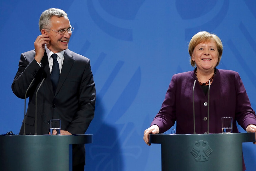 In front of a blue background bearing the German Chancellery logo, Angela Merkel and Jens Stoltenberg smile behind lecterns.