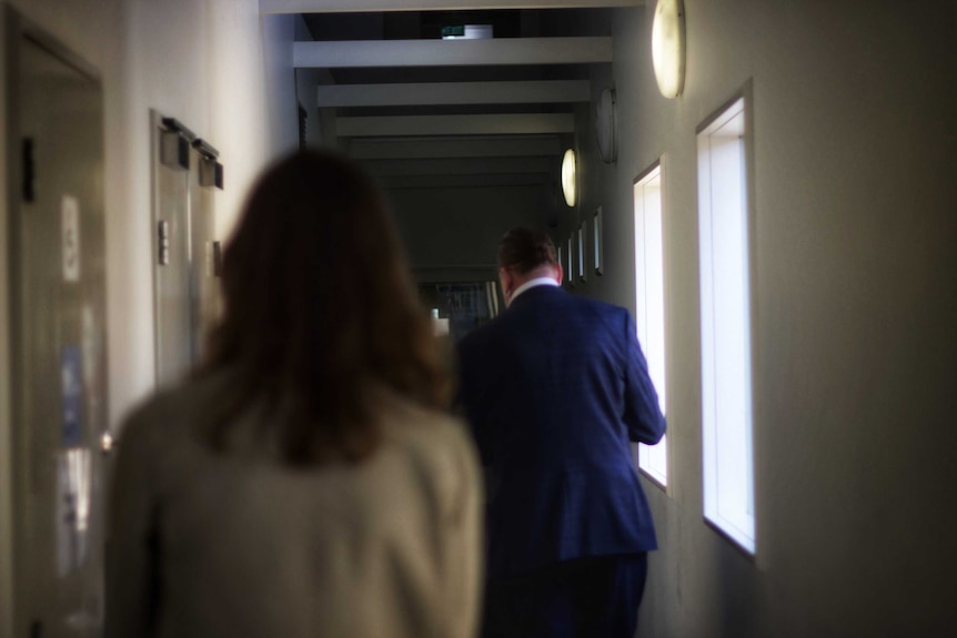 Two people walk down a hallway, there are a number of doors on either side.