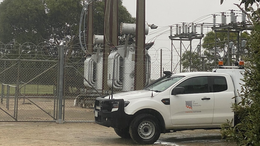 A white car parked outside a power station with electrical infrastructure in the background
