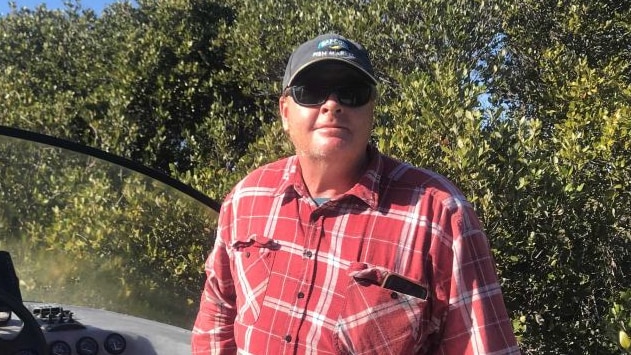 A man ina checked shirt and cap smiling with trees behind