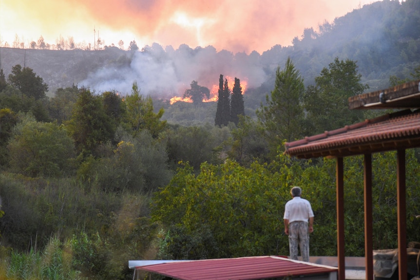 A man in a white shirt stands at front of house and watches fire burning in distance through wooded area