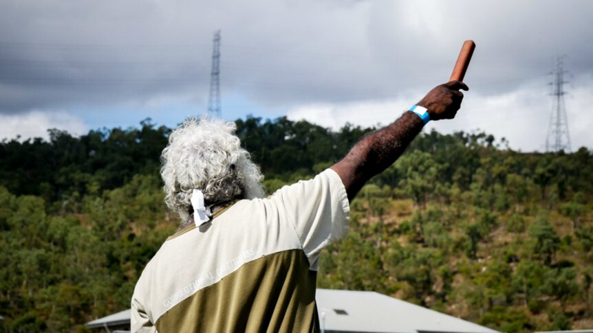 An Indigenous man with white hair, raising his arm. Photo taken from behind.