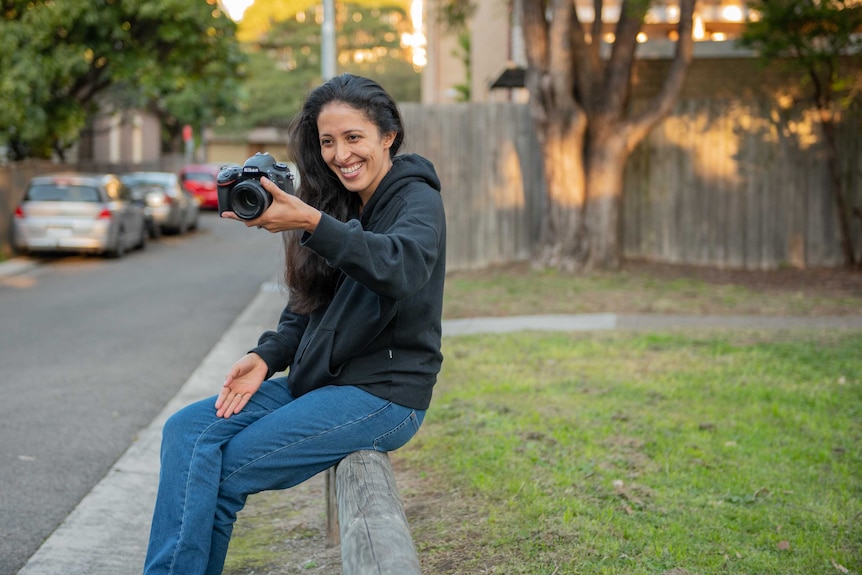A smiling woman with dark hair holds up a camera as she sits on a small fence near a park.