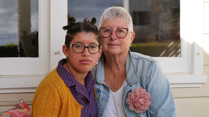 A woman and her adult daughter sit together on their front steps, their heads close and not smiling.