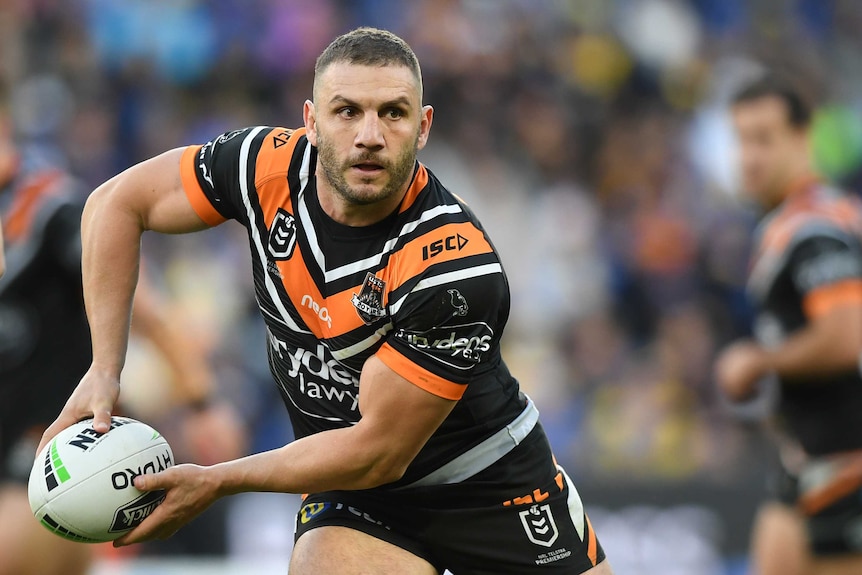 A male NRL player holds the ball in both hands as he looks to his left in preparation to pass the ball.