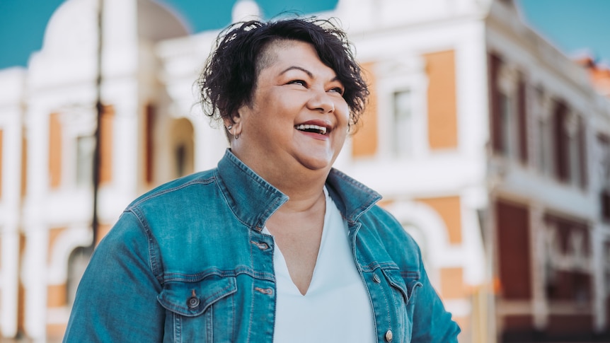 A woman wearing a white dress and denim jacket smiling in front of a heritage building.  