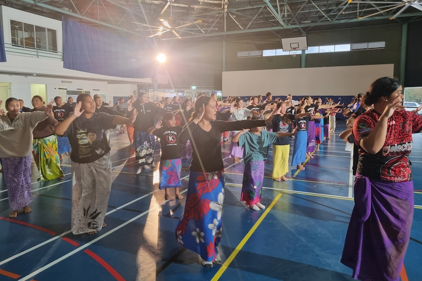 Rows of Samoan children are dancing Samoan Siva in large school hall with blue floors. 