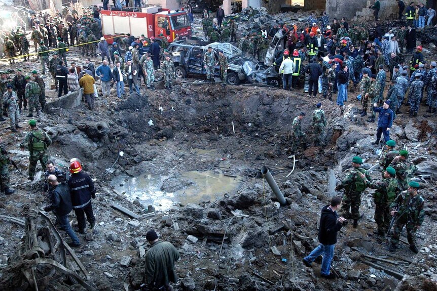 A huge crater in a street surrounded my men in military uniforms, a burned out car and a fire engine.