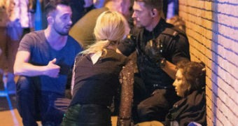 People huddle together around a girl sitting against a brick wall, covered in others' coats.