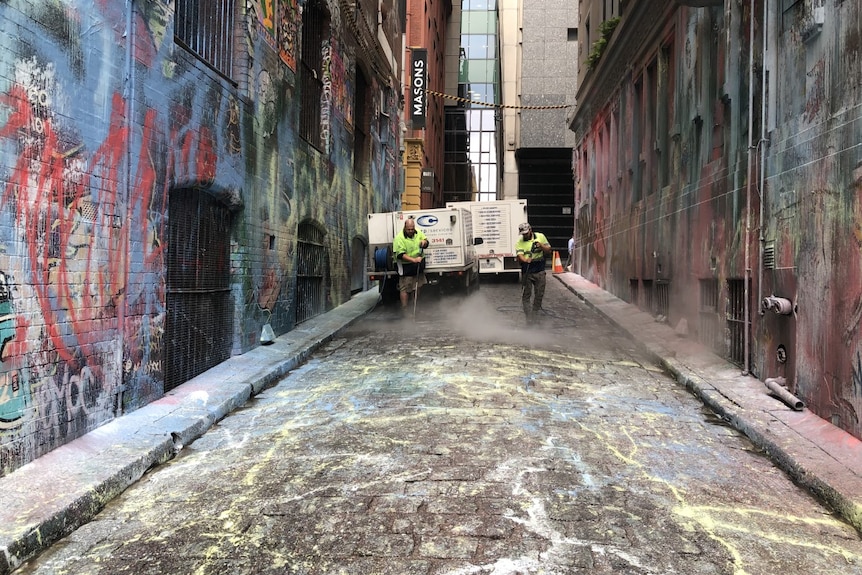 Two street cleaners wearing high vis clean a cobble stone laneway of spray paint.