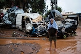 A barefoot man stands in front of a pile of wrecked vehicles.