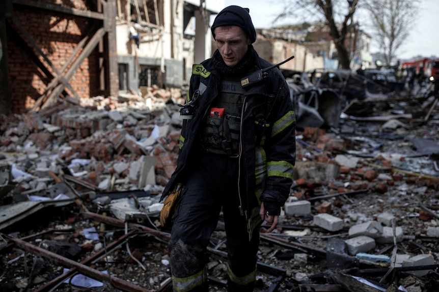 A stunned looking man in firefighter gear walking through rubble 