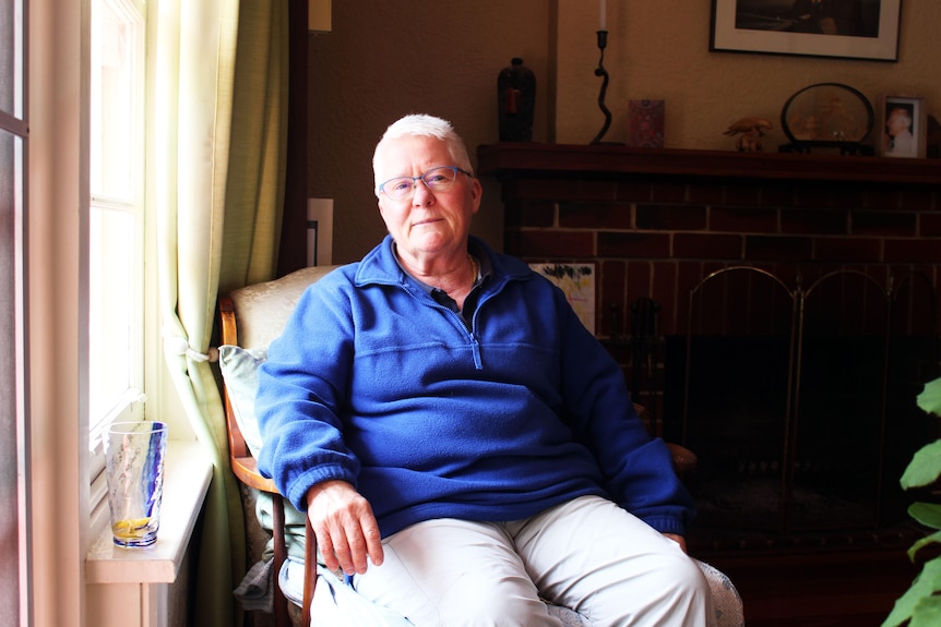 Older woman with white hair and blue jumper sits next to a window 