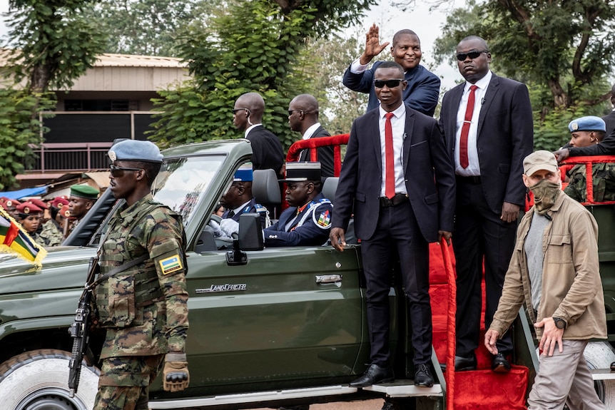 A man waves from the back of an open truck, while soldiers walk nearby and two security guards in suits