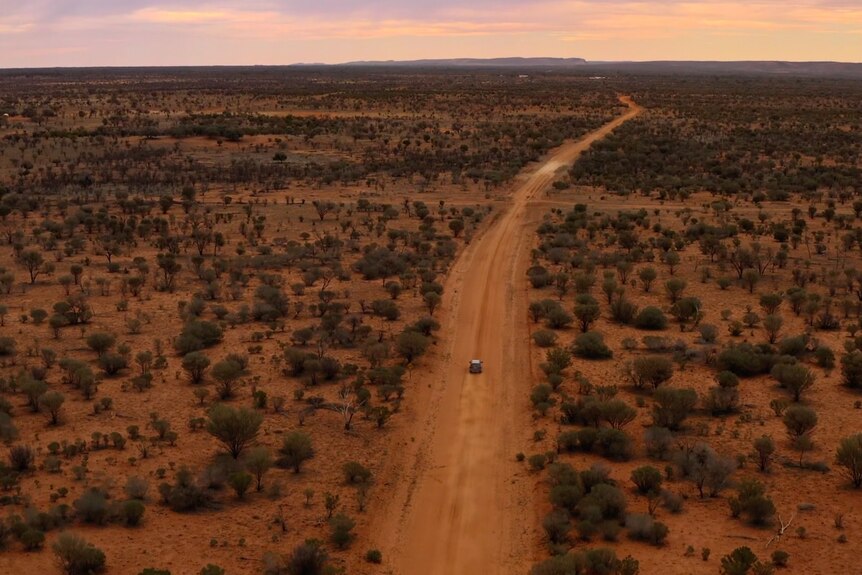 Aerial shot of a car driving down a red dirt road in the bush.