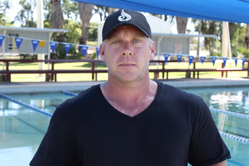 Hombre con sombrero negro y camisa parado frente a una piscina. 